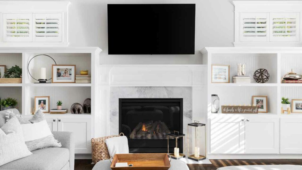 TV above fireplace. White fireplace surround. Book shelf to one side and picture frames the other side.