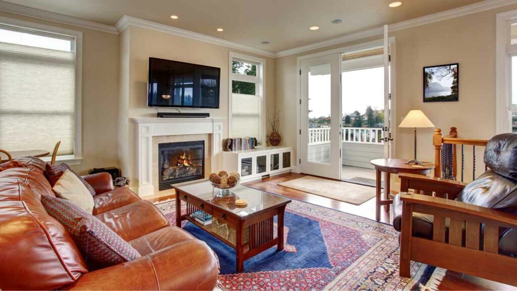 Wood burning fireplace. TV on wall above. Brown leather sofa. Red rug carpet. Small table in front of the fire. Book sshelving to one side. Large open doors with a view.