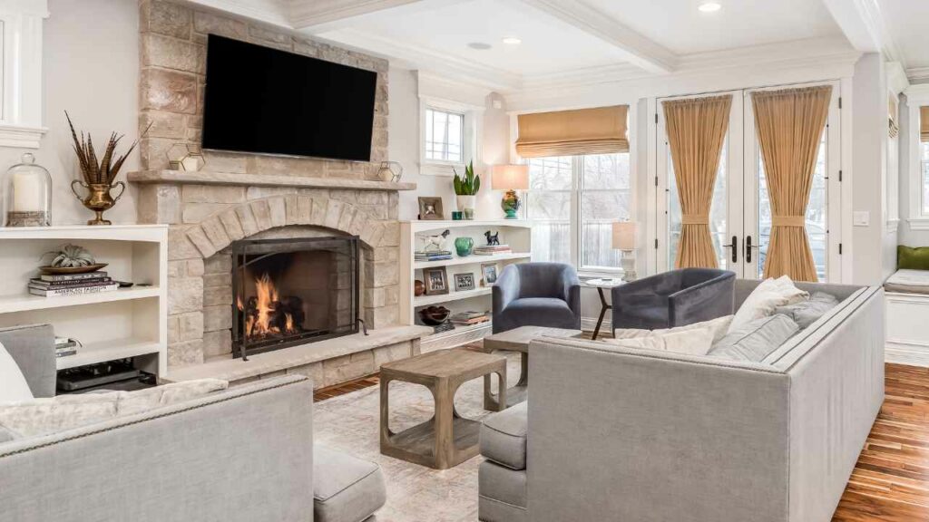 TV above fireplace, stone fireplace surround. Cream/Grey sofa in front of a wood burning fireplace.