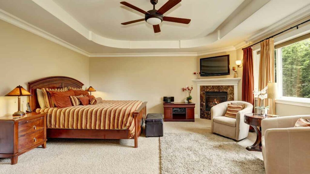 Corner fireplace in the bedroom, tv above. rug on the floor. ceiling fan. armchair next to the fireplace.