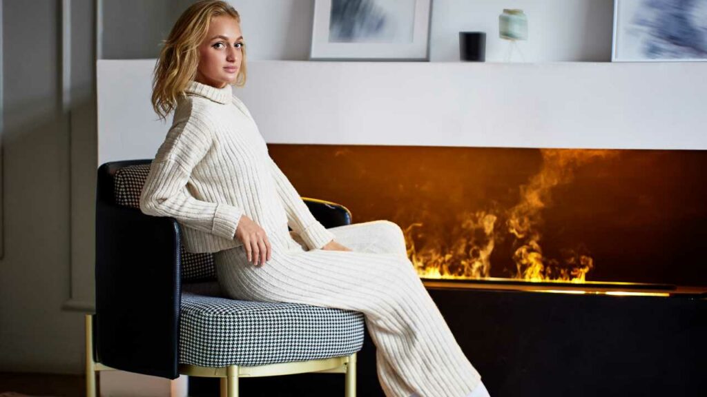 Electric Vapor fireplace. Young woman keeping warm in front sitting on chair.. Modern design. Photograph on mantel.