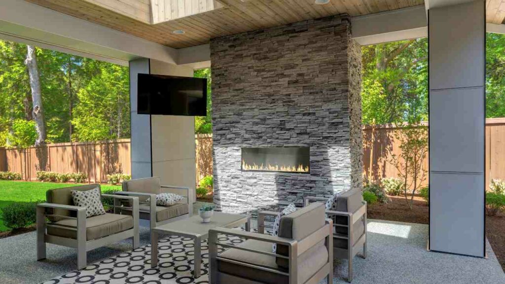 Outdoor Gas fireplace set in brick fireplace. Chairs and tables in front. In pergola. TV set to the side.