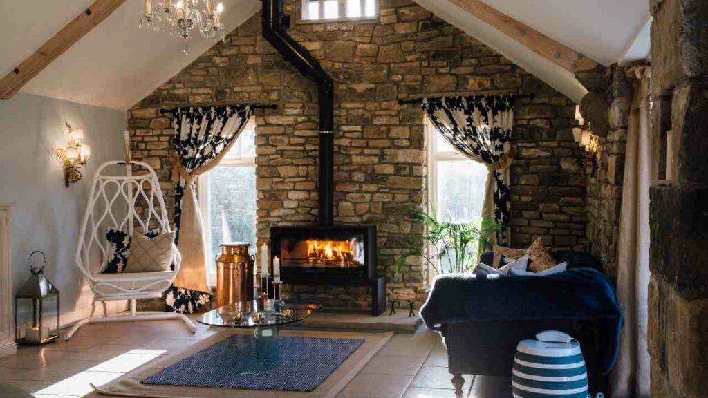 wood burning fireplace with stone fire surround. Hearth in front in slab concrete with tiled floor and blue rug. Blue sofa to one side and windows either side of the fireplace.
