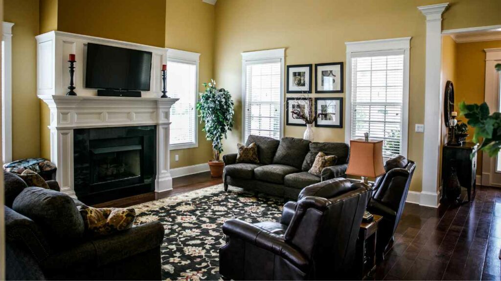 TV above fireplace with candles either side of the TV. White marble fireplace with rug in front. 2 sofas either side and leather chair in front of the TV.