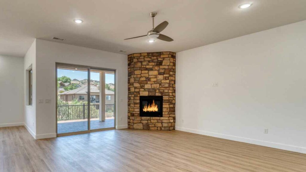 rustic stone corner fireplace. wood flooring. Empty room with ;large windows with a view outside.