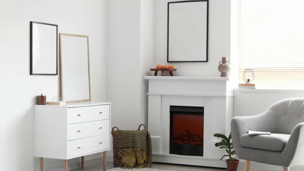 Corner fireplace in white, with white walls and white set of draws. Grey lounge chair. Next to small window with blinds drawn. mirror above the fiureplace.