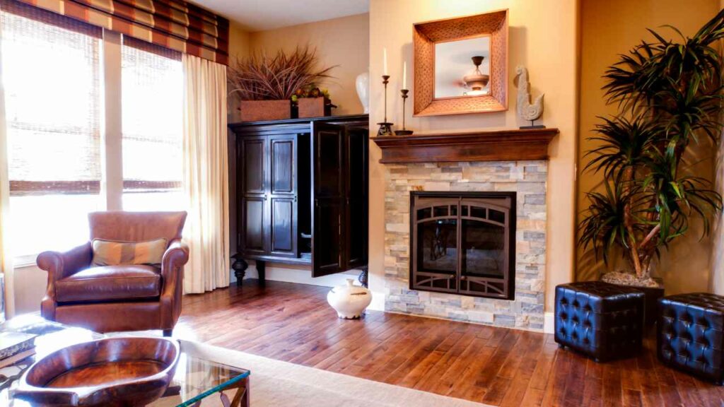 Fireplace with TV in cabinet in alcove. Mustard colored walls with wood flooring. large windows with curtains. leather chair to the side of the fireplace.