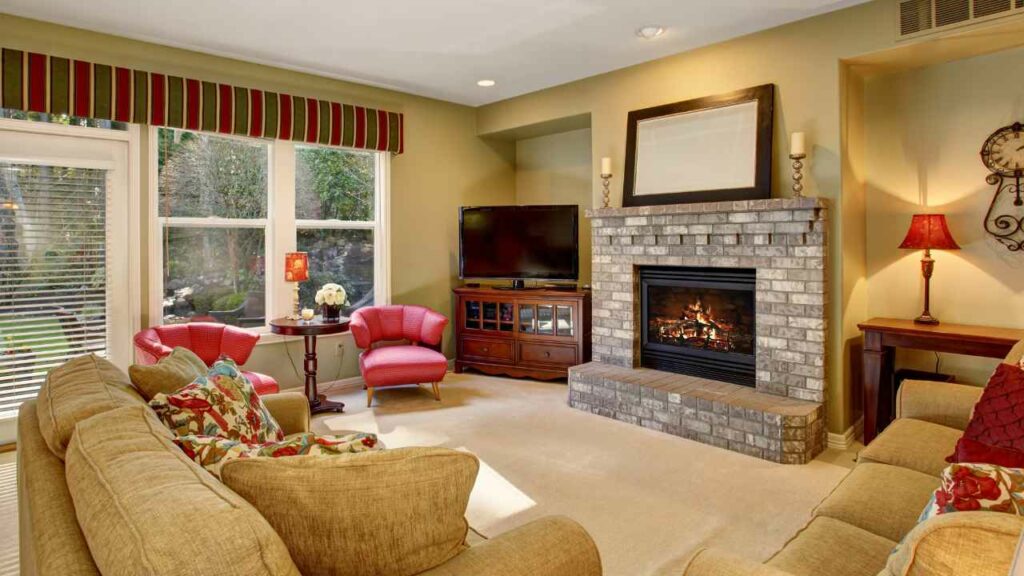 brick fireplace surround and hearth. mirror above. two mustard cloured sofas. Tv in the orner next to a large window and glass door.