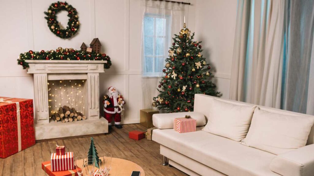 Cream faux fireplace with logs inside. christmas decorations. Fairy lights hanging down from the mantel. Wood flooring. Small table and white sofa in fromnt of the fireplace.