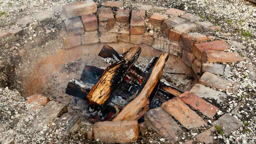 Rustic sunken fire pit. chared logs in the centre. red brick for the walls stacked.