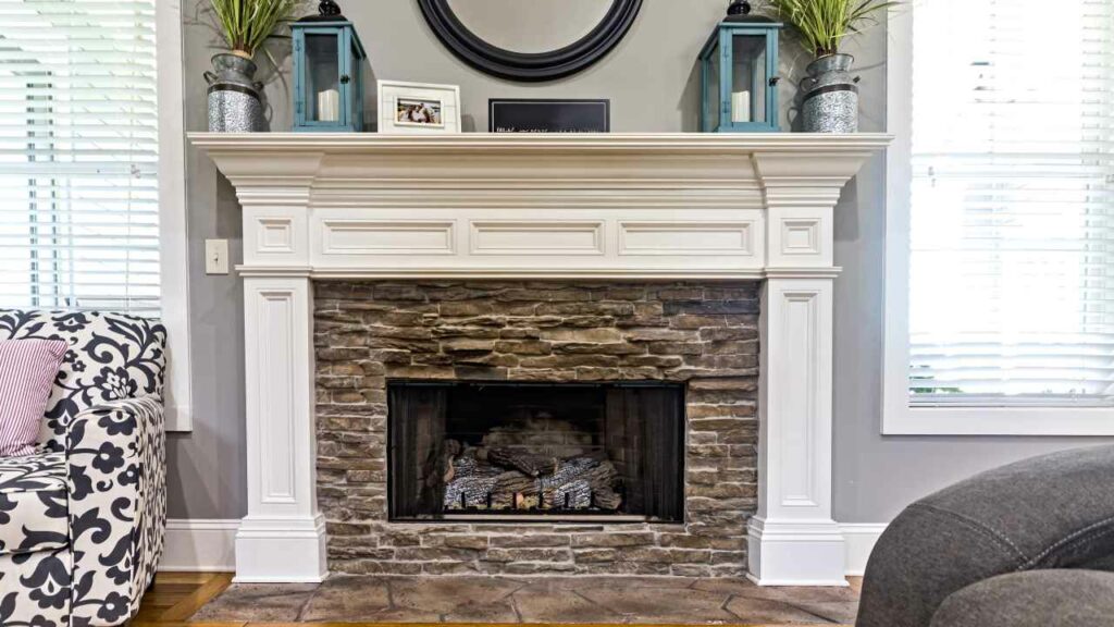 Fireplace with stone vaneer fireplace surround. white mantel. tiled hearth. round mirror above. ornaments on the mantel.