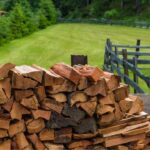 large pile of wood logs. In garden with green lawn and fence.