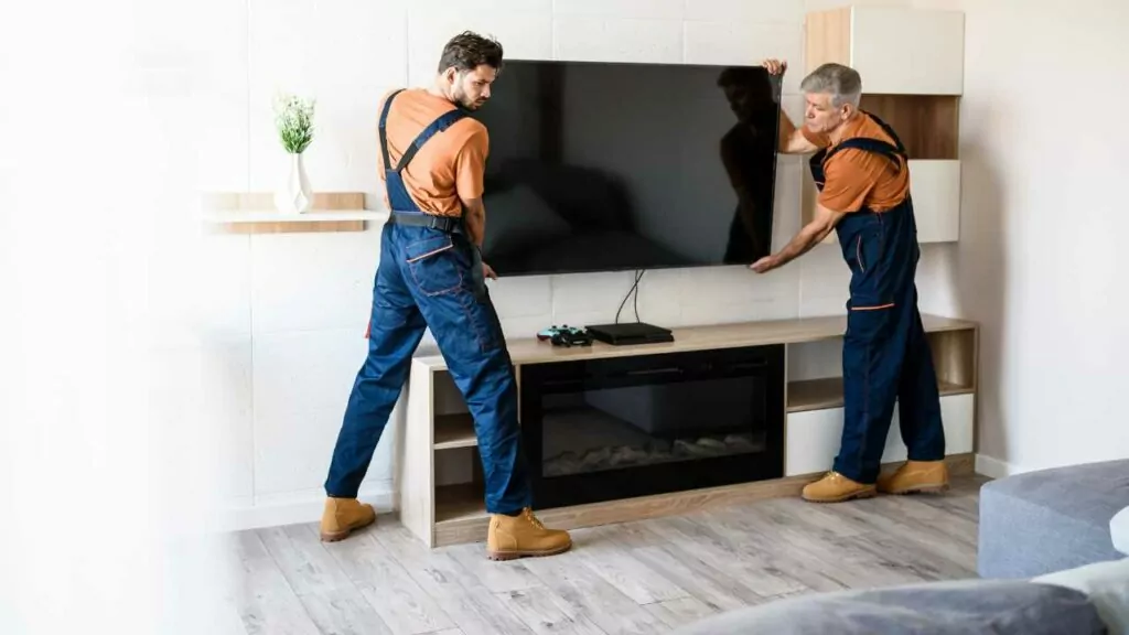 2 Men Hanging TV on a wall. Fireplace below with mantel. Cable box on the mantel.