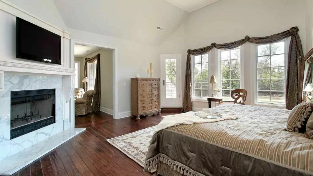 Bedroom |White Marble Fireplace with TV above. Wood Flooring. Large Windowns and large bed.