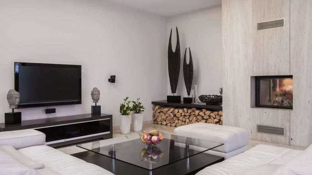 Tv place on wall with the fireplace placed on another wall to the side. Wood log storage next to the fireplace with decorations above. Corner sofa in cream and tiled flooring.